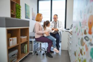 A girl is with her mom while being examined by a male pediatrician during her wellness exam.