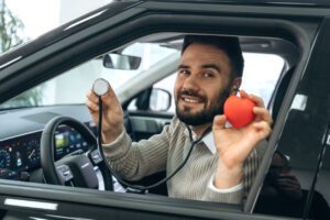 A man is sitting in his car, holding a stethoscope and a small red heart model to illustrate a DOT physical exam.