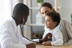 A male family physician and pediatrician listens to a child's lung and heart sounds in the presence of the mother.