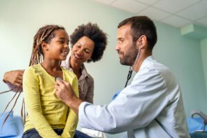 A cheerful girl with her mother and a male family medicine doctor, wearing a stethoscope, during her routine health screenings.